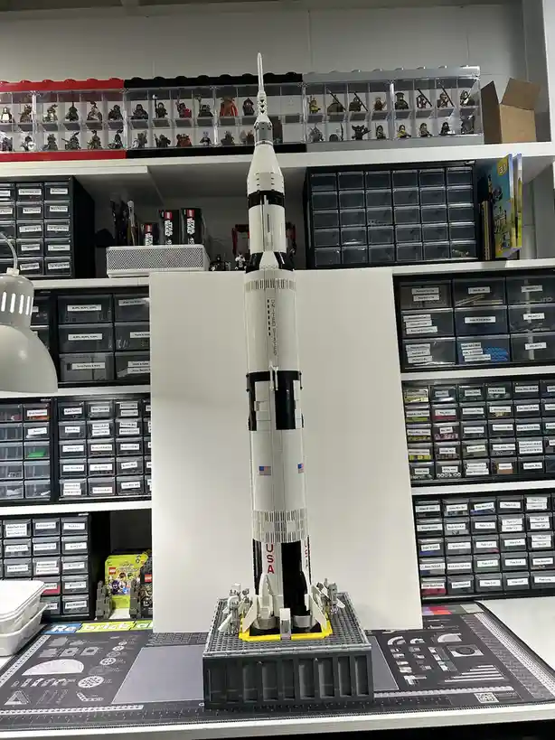 A large, detailed LEGO model of a Saturn V rocket stands on a build mat in front of organized LEGO storage shelves. The…