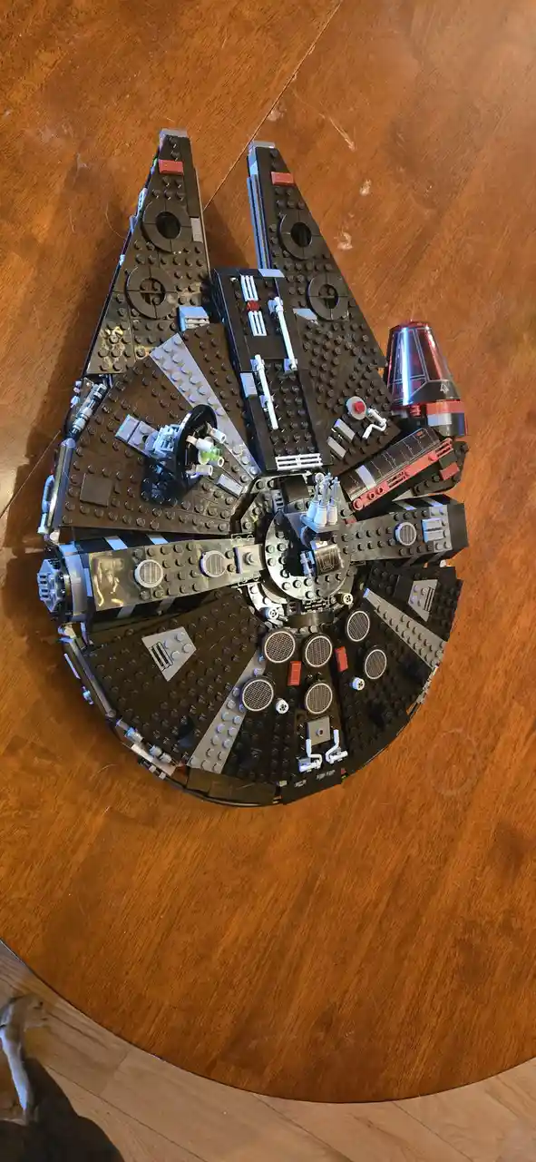 A top-down photo shows a large, partially assembled LEGO Millennium Falcon model resting on a wooden table. The intricate…