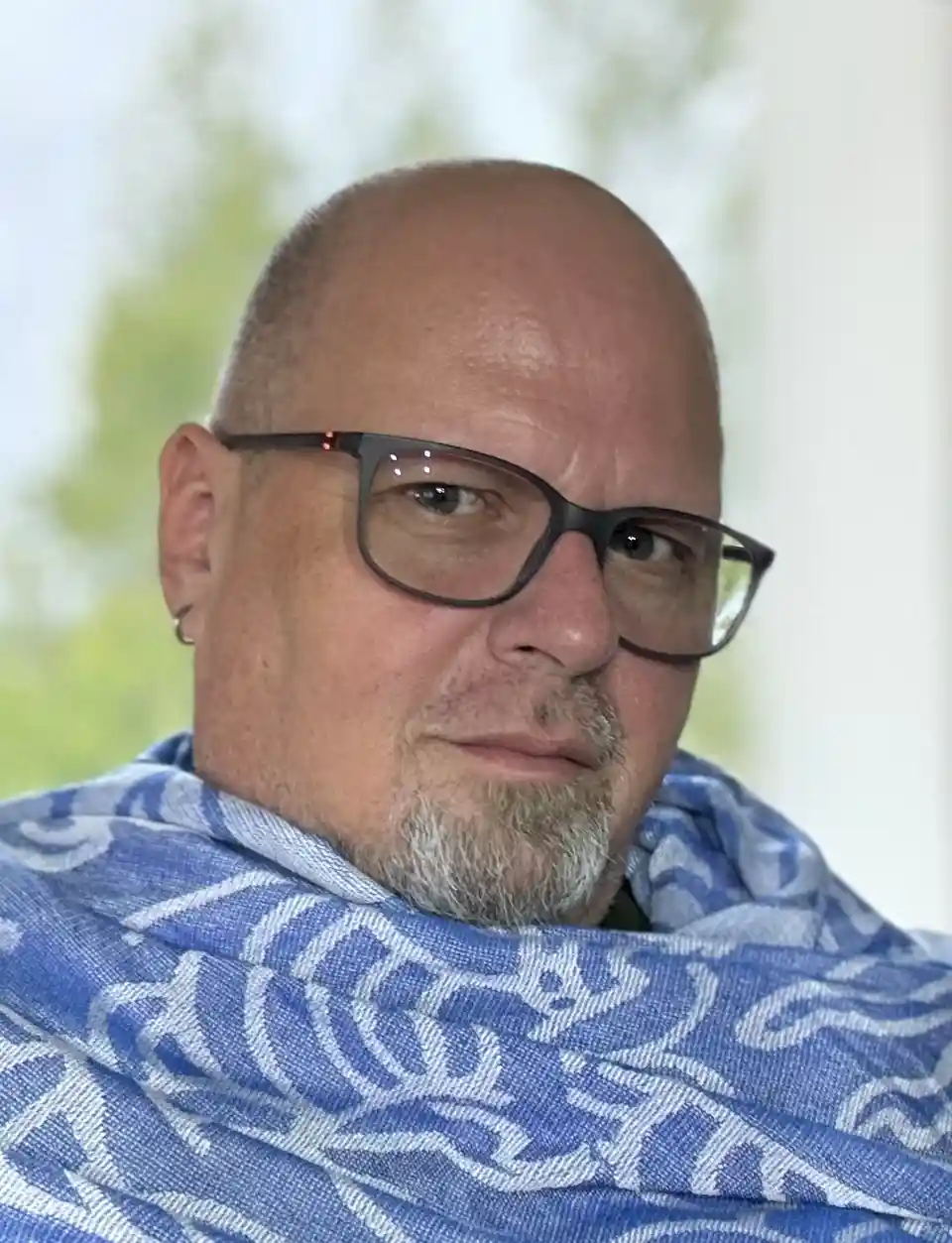 A close-up photo of a bald man with glasses and a goatee, wearing a blue and white patterned scarf, looking directly at the…