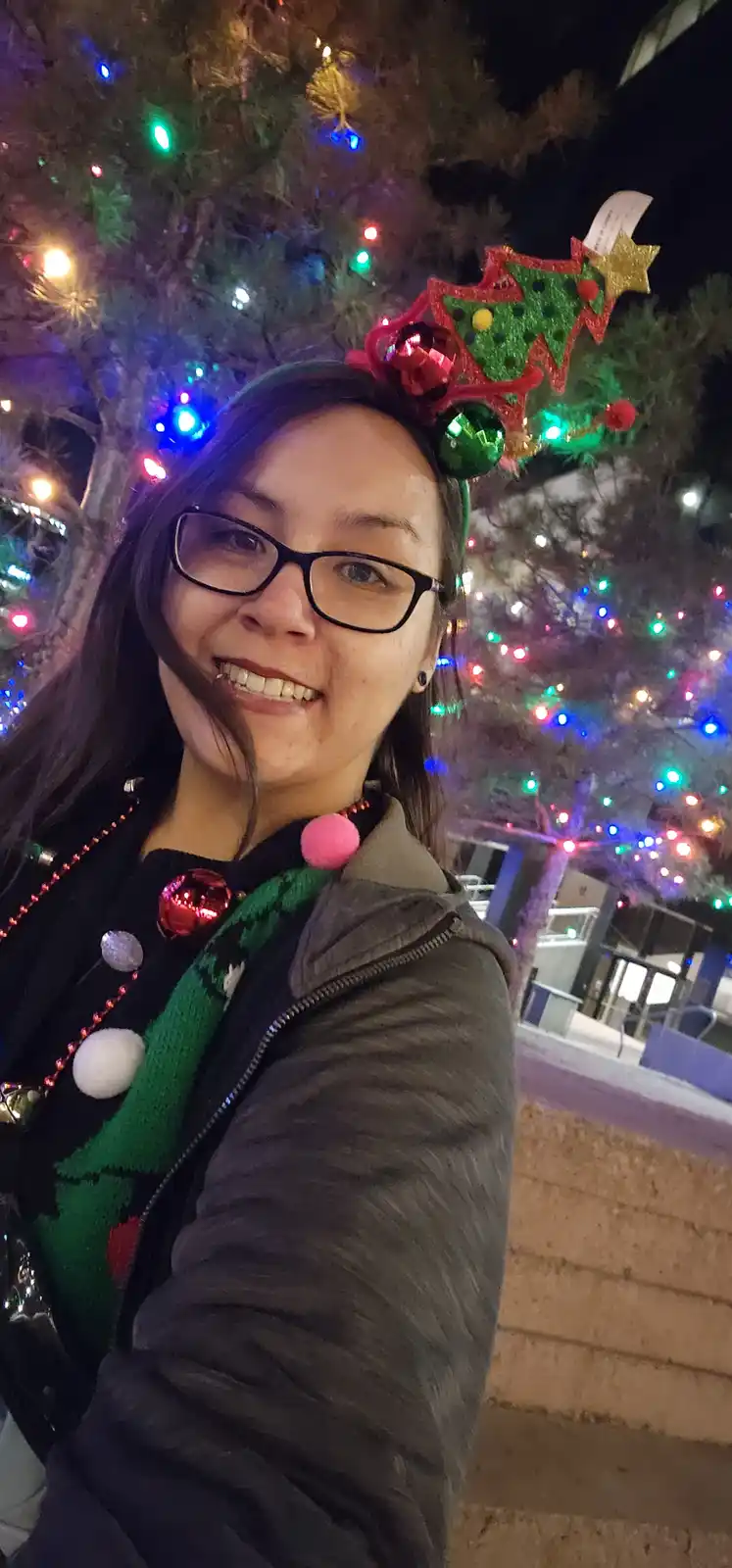 A person wearing glasses and a festive Christmas tree-themed headband and sweater smiles for a selfie in front of a tree…
