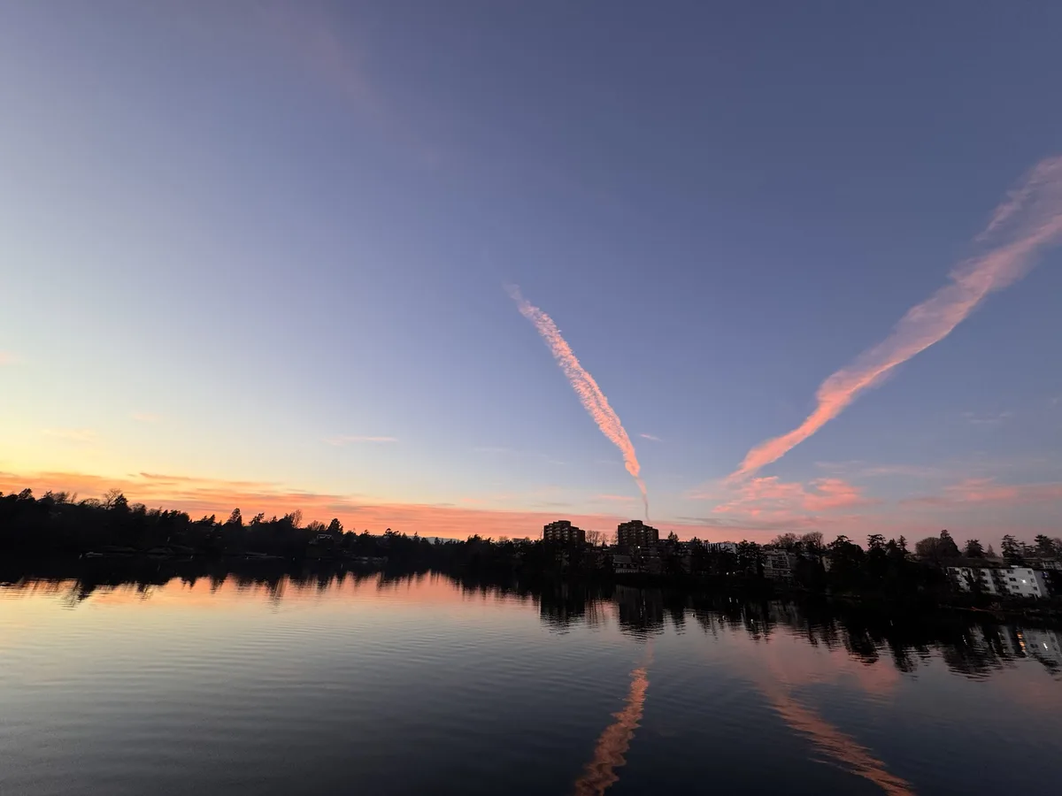 A serene sunset over a calm lake with city buildings silhouetted on the shore, featuring two long, thin, pink-tinged cloud…