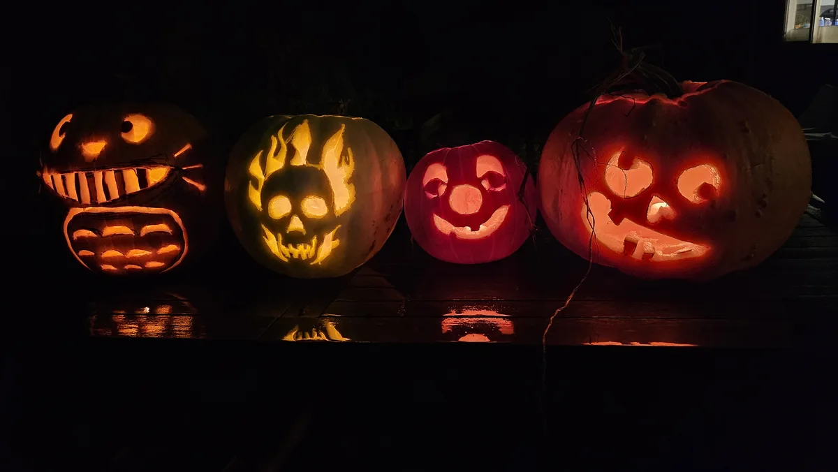 Four carved pumpkins with glowing faces sit in a row on a dark wooden surface at night. From left to right: a pumpkin with a…