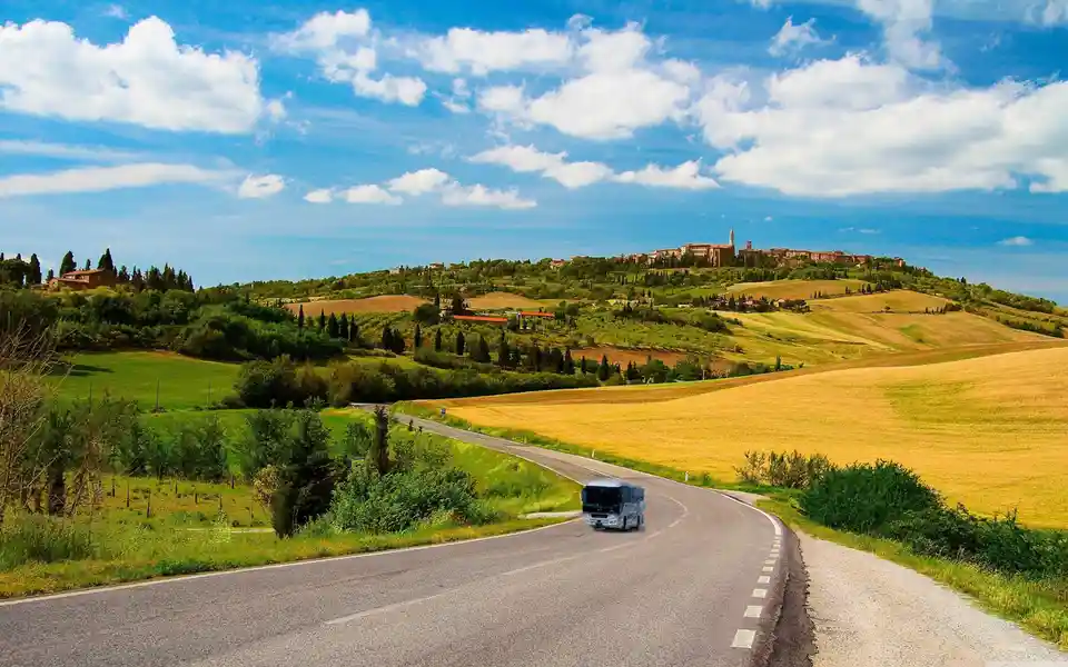 A scenic photo of a winding road through rolling green and golden hills under a bright blue sky with white clouds, leading…