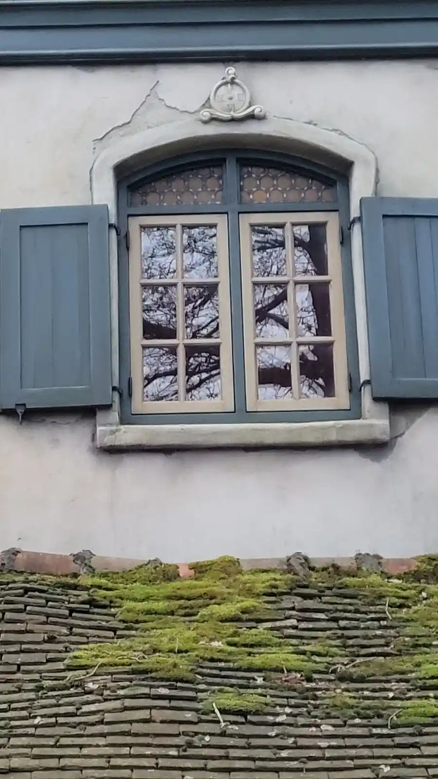 A close-up of an old European-style window with blue shutters and a decorative stone clock above it, set in a weathered…