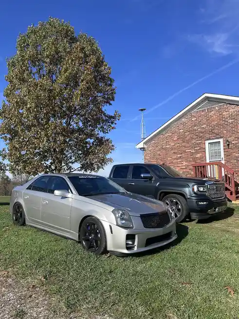 Two vehicles are parked on a grassy lawn in front of a brick house under a clear blue sky. A silver Cadillac CTS-V with…