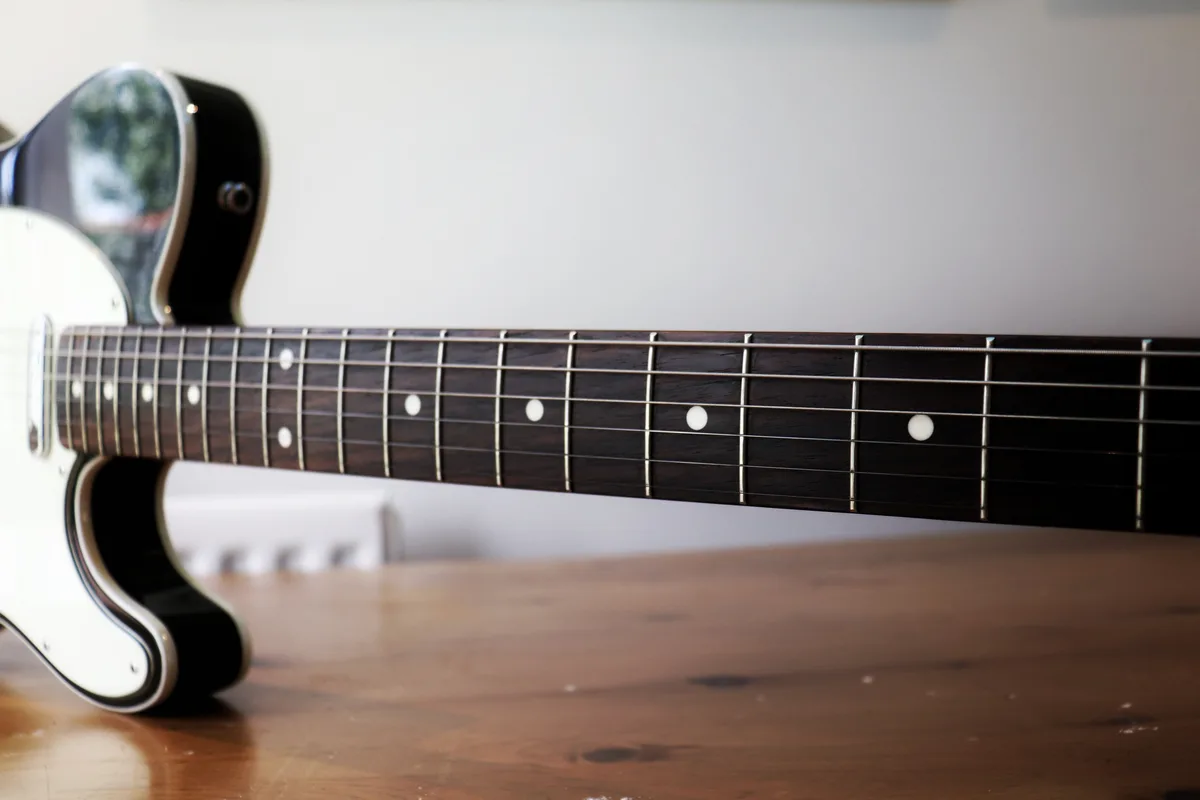 A close-up photo of an electric guitar's fretboard resting on a wooden surface, with the body partially visible. The image…