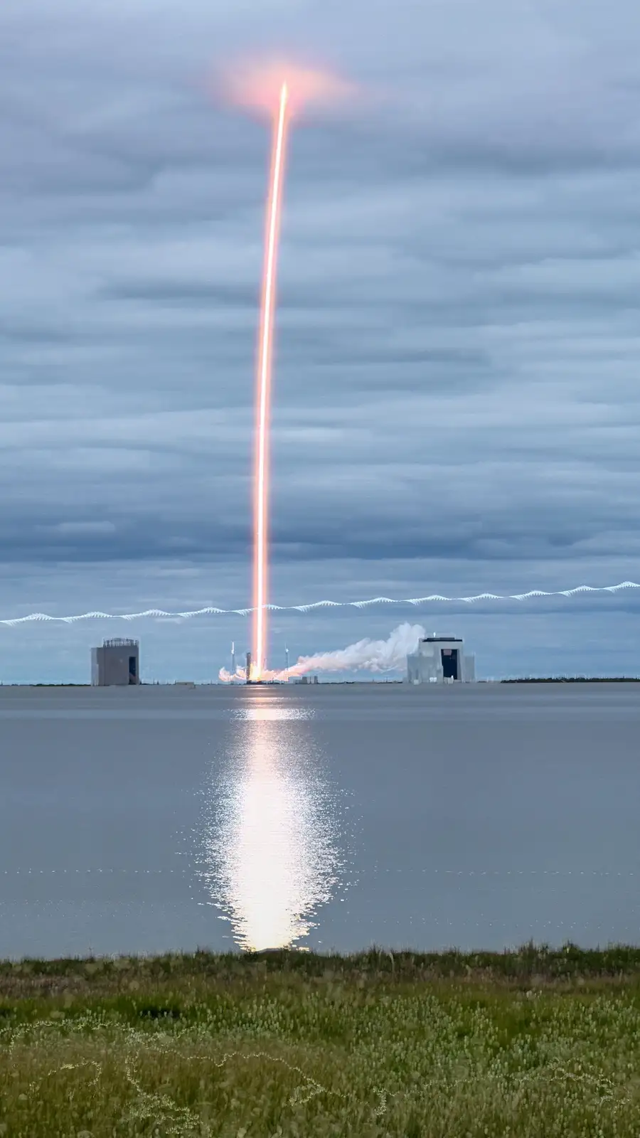 A rocket launches vertically over water, leaving a bright, fiery trail that reflects on the surface. The sky is overcast…