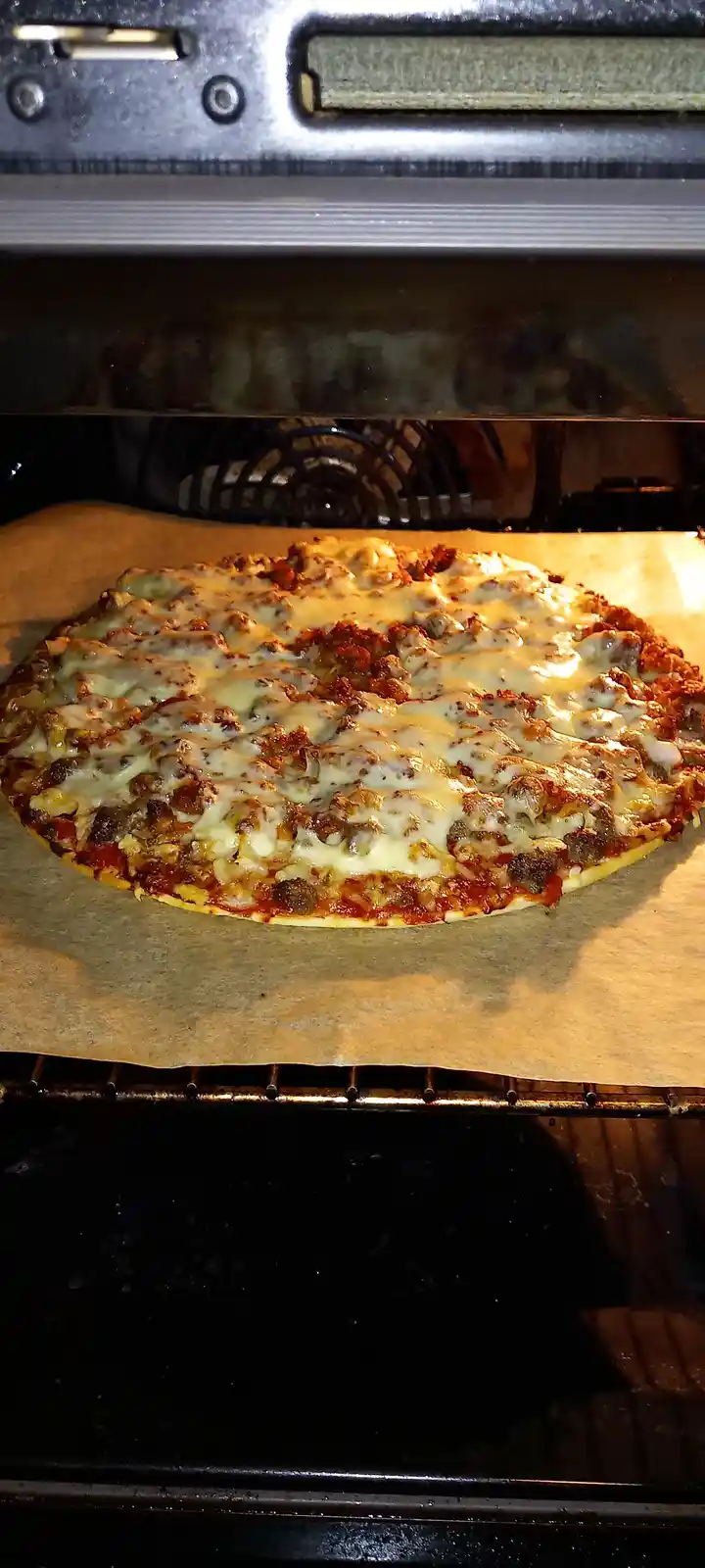 A close-up photo shows a round pizza with melted cheese and meat toppings baking inside an oven on parchment paper. The text…