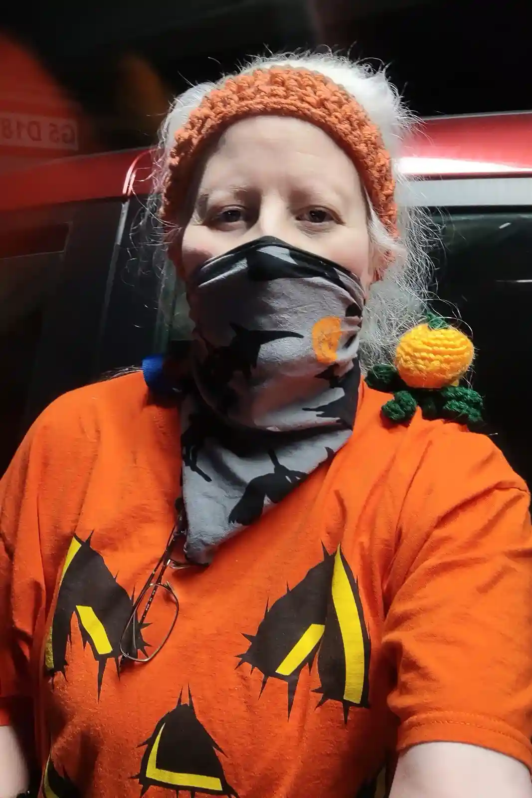 A person with white hair wears an orange knit hat, a gray face covering with black bat and pumpkin patterns, and an orange…