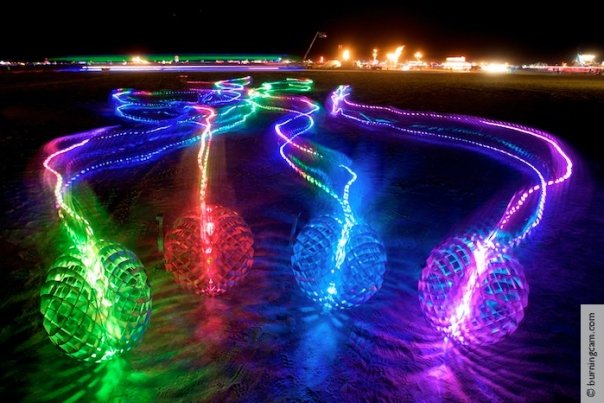 A long-exposure night photo shows four large, illuminated, grid-patterned spheres on sand, each emitting colorful light…