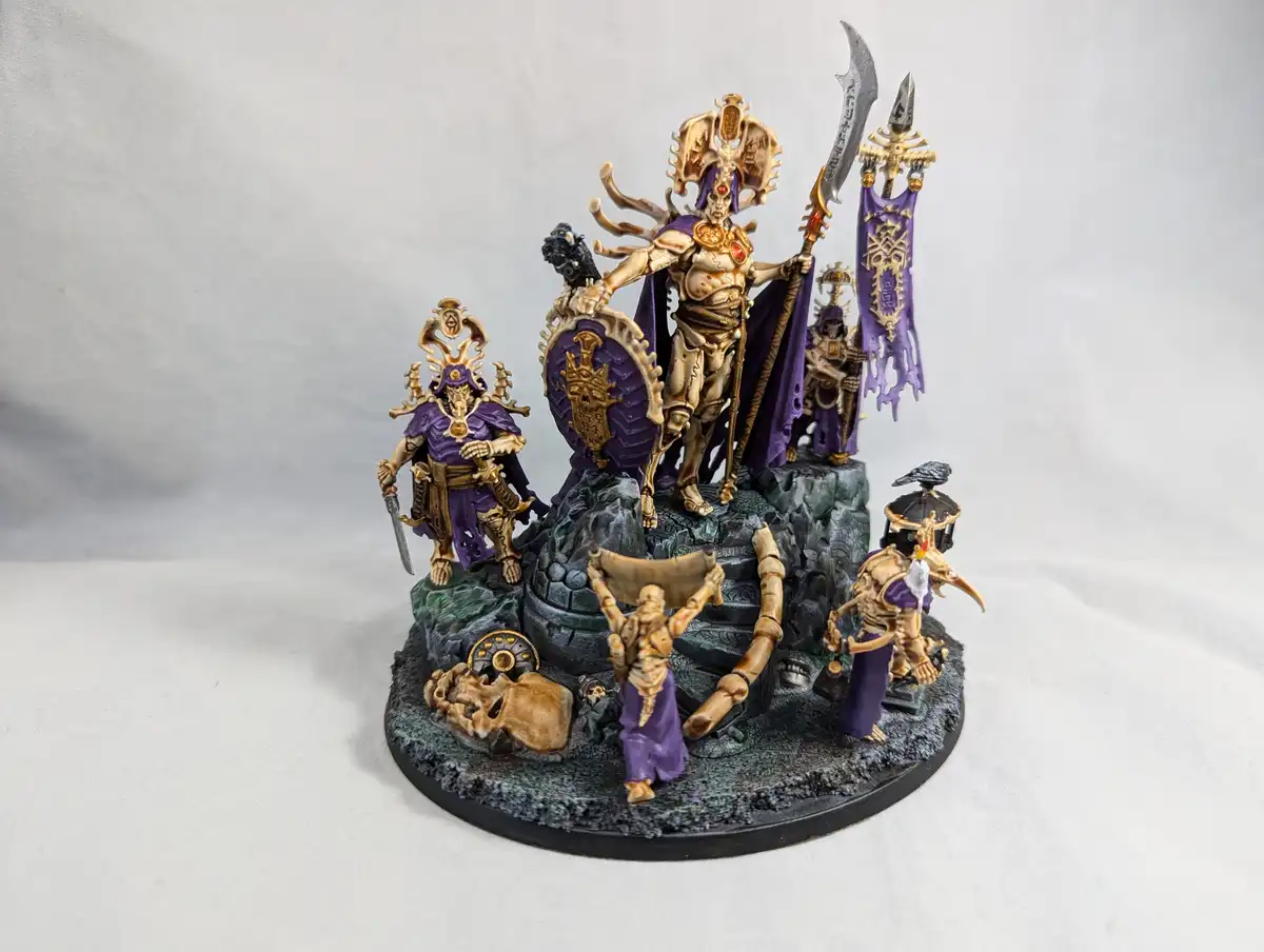 A painted miniature wargaming diorama featuring a central skeletal warlord with a purple cloak and ornate golden armor…
