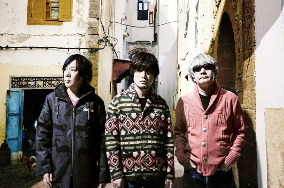 three men stand in a narrow, sunlit alleyway with weathered buildings and a blue door in the background. The man on the left…