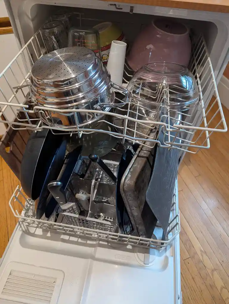 A dishwasher with both upper and lower racks filled to overflowing with dirty dishes including pots, pans, glassware, bowls…