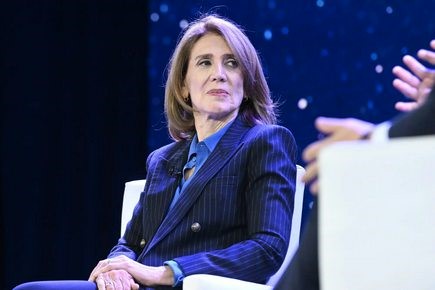 A woman in a dark pinstripe suit sits in a white chair, looking slightly confused or disengaged while a person's hands…