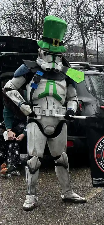 A person in a Star Wars clone trooper costume stands outdoors in the rain, wearing a large green leprechaun top hat with a…