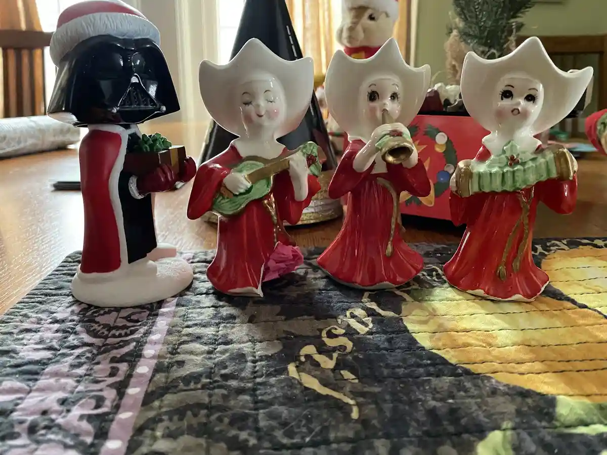 A photo shows a Darth Vader figurine dressed in a Santa hat and red-and-white outfit, holding a gift box, standing next to…