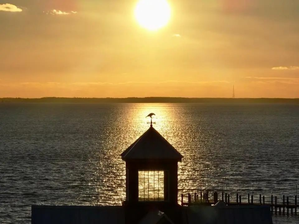 A serene sunset over water with a weather vane atop a small structure in the foreground. The sun is low, casting golden…