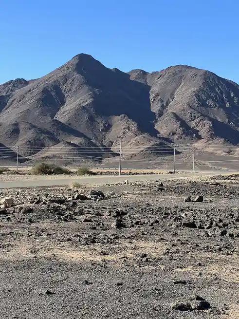 A barren, rocky desert landscape with a paved road running horizontally, flanked by utility poles and wires, set against a…
