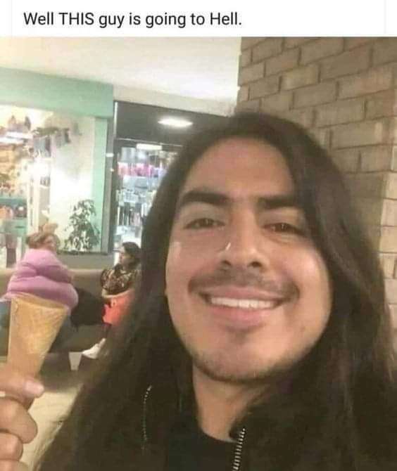 A man with long dark hair and a wide, toothy grin holds an ice cream cone with purple swirls. He is taking a selfie in front…