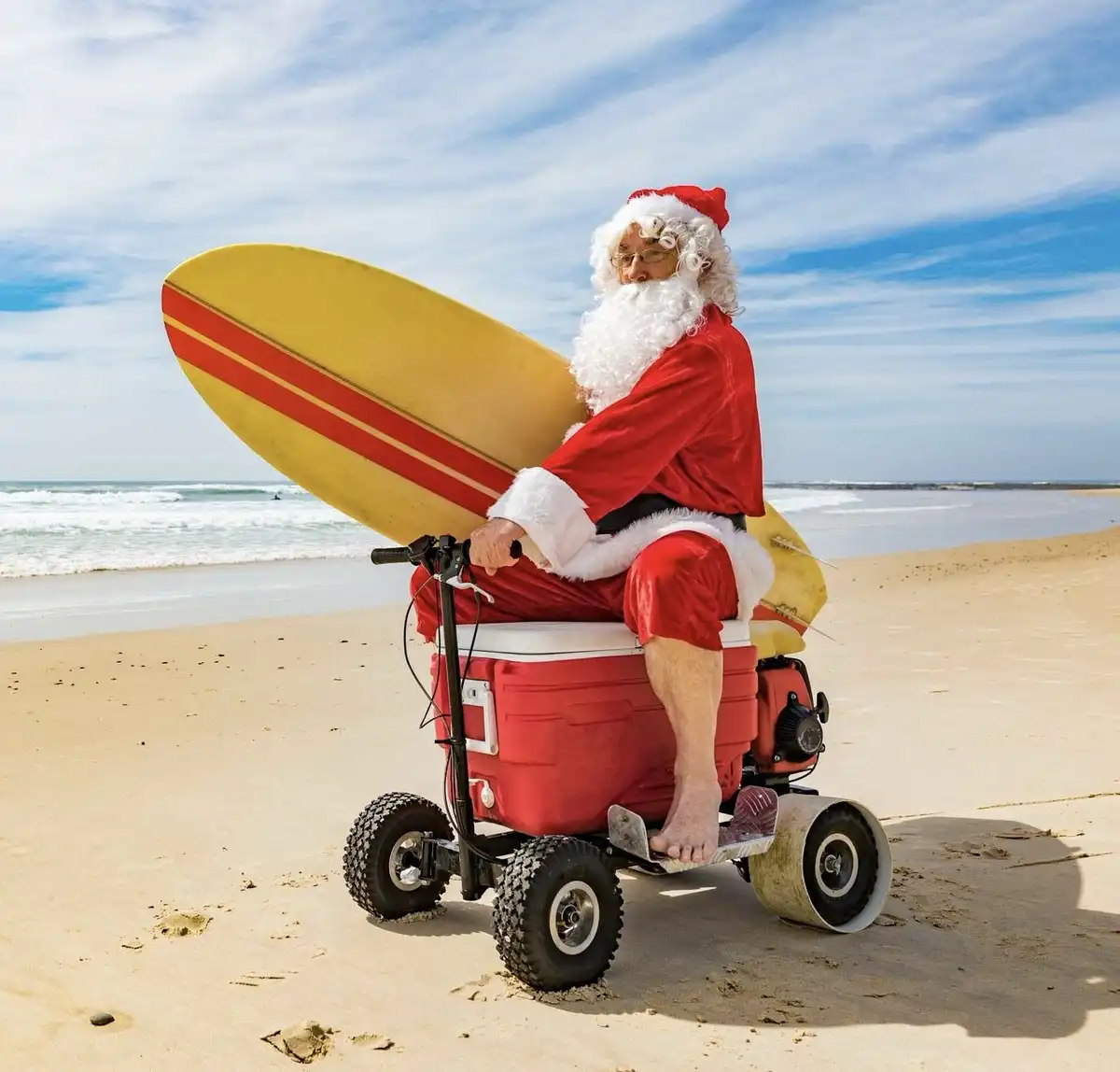A man dressed as Santa Claus, complete with red suit, white beard, and hat, is seated on a red cooler modified into a…