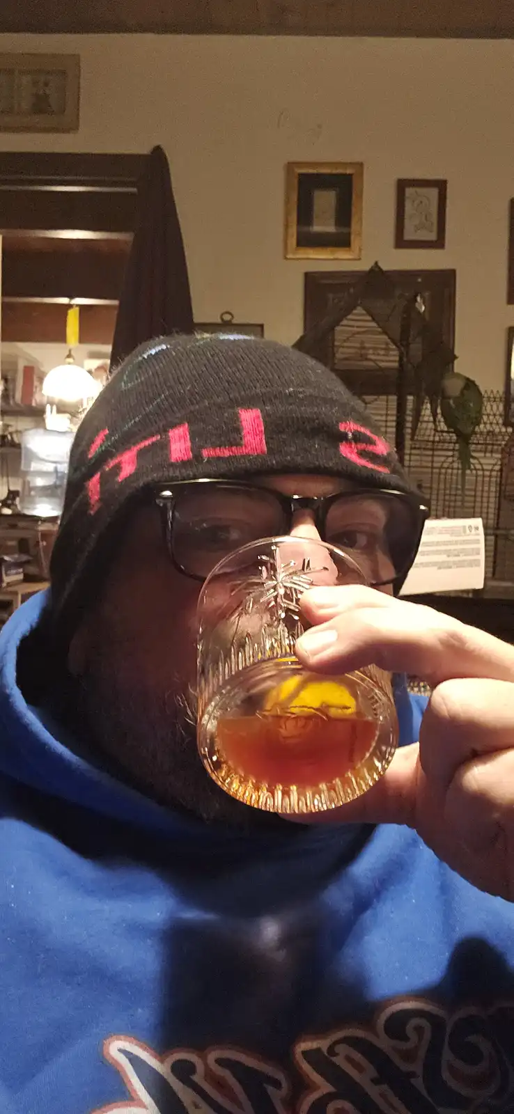 A man wearing a black beanie with pink 'FILS' text, glasses, and a blue hoodie is holding a glass of amber liquid with a…