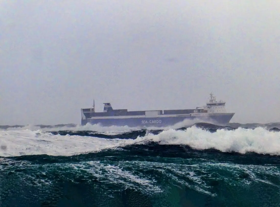 A large cargo ship named 'SEA CARGO' sails through rough, choppy ocean waves under an overcast sky. The vessel is partially…