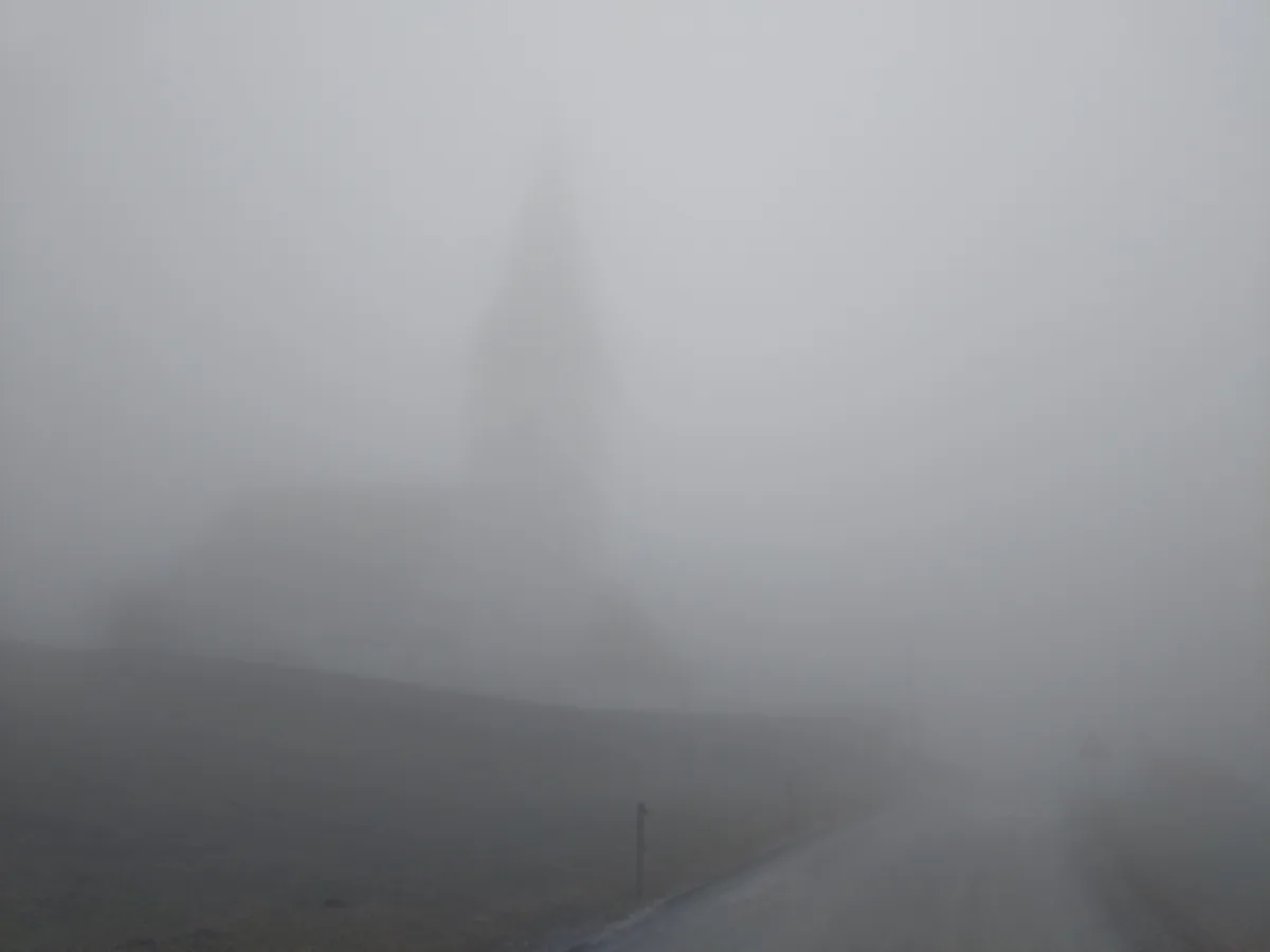 A foggy rural road leads toward a barely visible church steeple shrouded in thick mist, with the word 'SOLAR' painted on the…