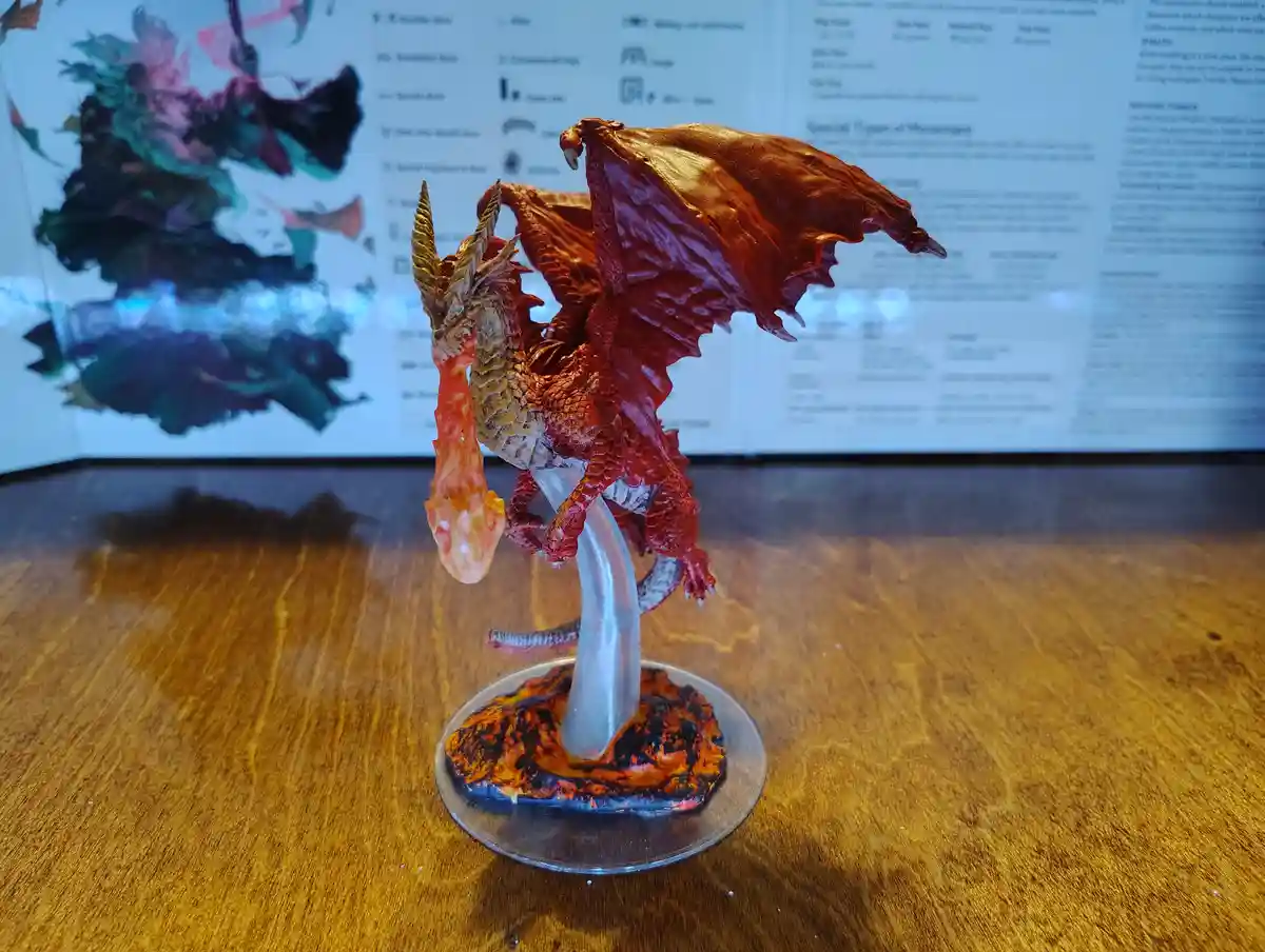 A painted miniature dragon figurine with red wings and a fiery breath is mounted on a clear plastic stand with a…