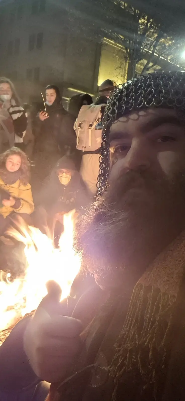 A man with a long beard and chainmail coif takes a selfie at night near a campfire, holding up a lit torch. Behind him…