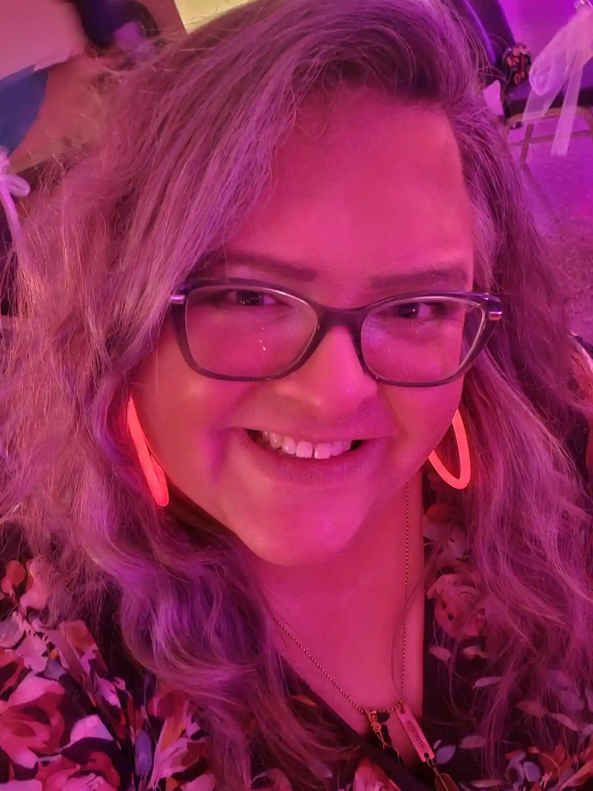 A close-up photo of a smiling woman with glasses and large hoop earrings under intense pink and purple lighting, likely at a…