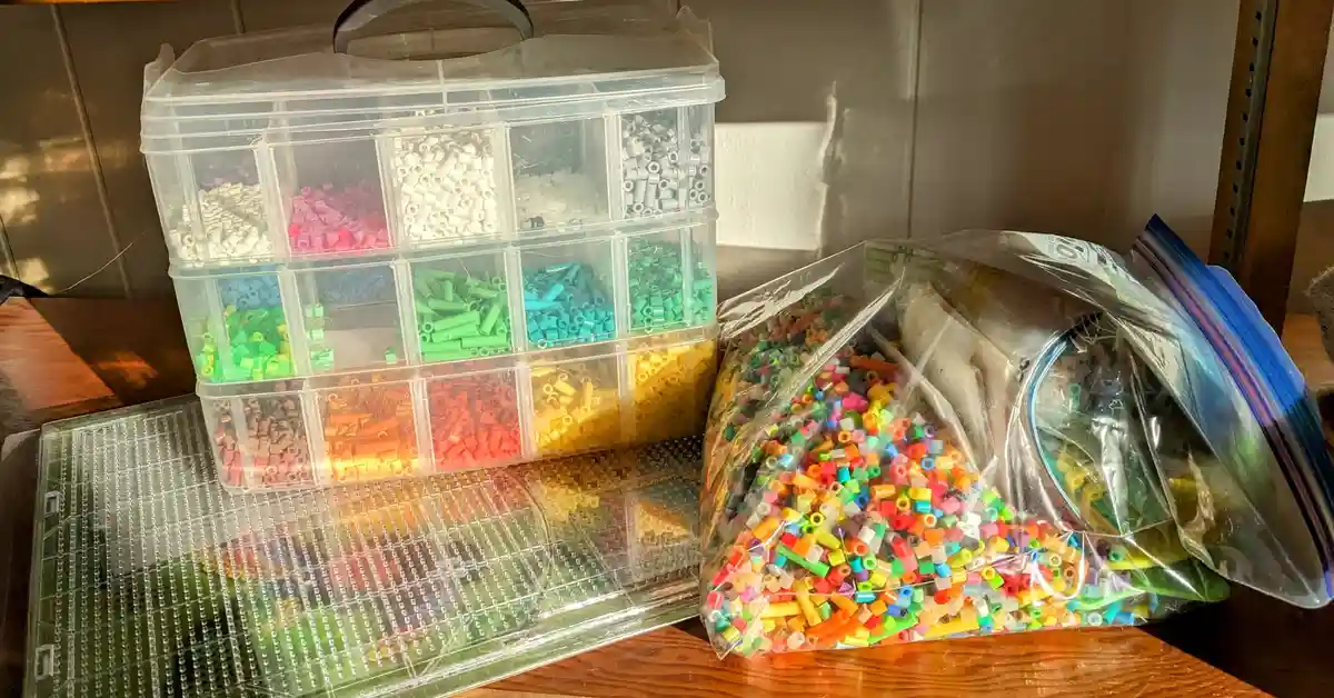 A clear plastic organizer with multiple compartments filled with sorted, colorful perler beads sits next to a large, messy…