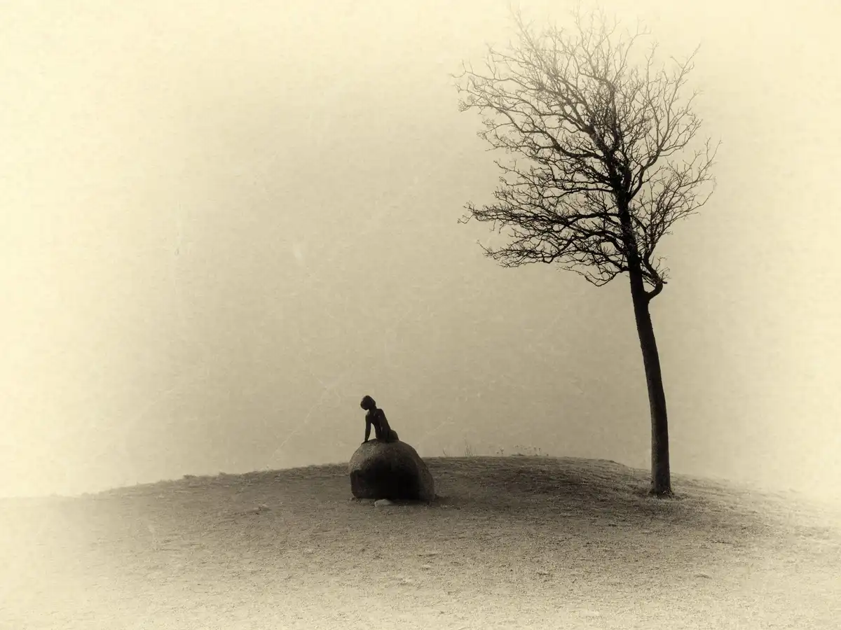 A sepia-toned, atmospheric image shows a small human figure sitting atop a large boulder on a barren hill, with a solitary…