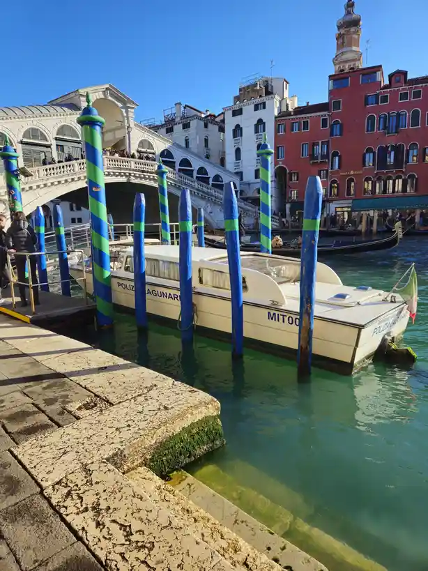 A white police boat labeled 'POLIZIA LAGUNARE' and 'MITOO' is docked in Venice's canal, surrounded by blue and green striped…