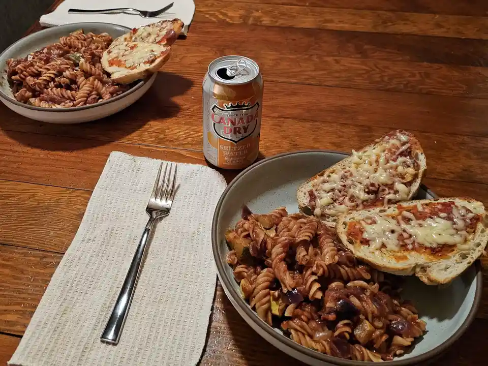 A wooden table set with two bowls of pasta with sauce and melted cheese on bread, a can of Canada Dry Mandarin Orange…