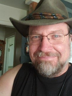 A middle-aged man with glasses and a goatee wears a brown cowboy hat with a beaded band, smiling slightly while looking at…