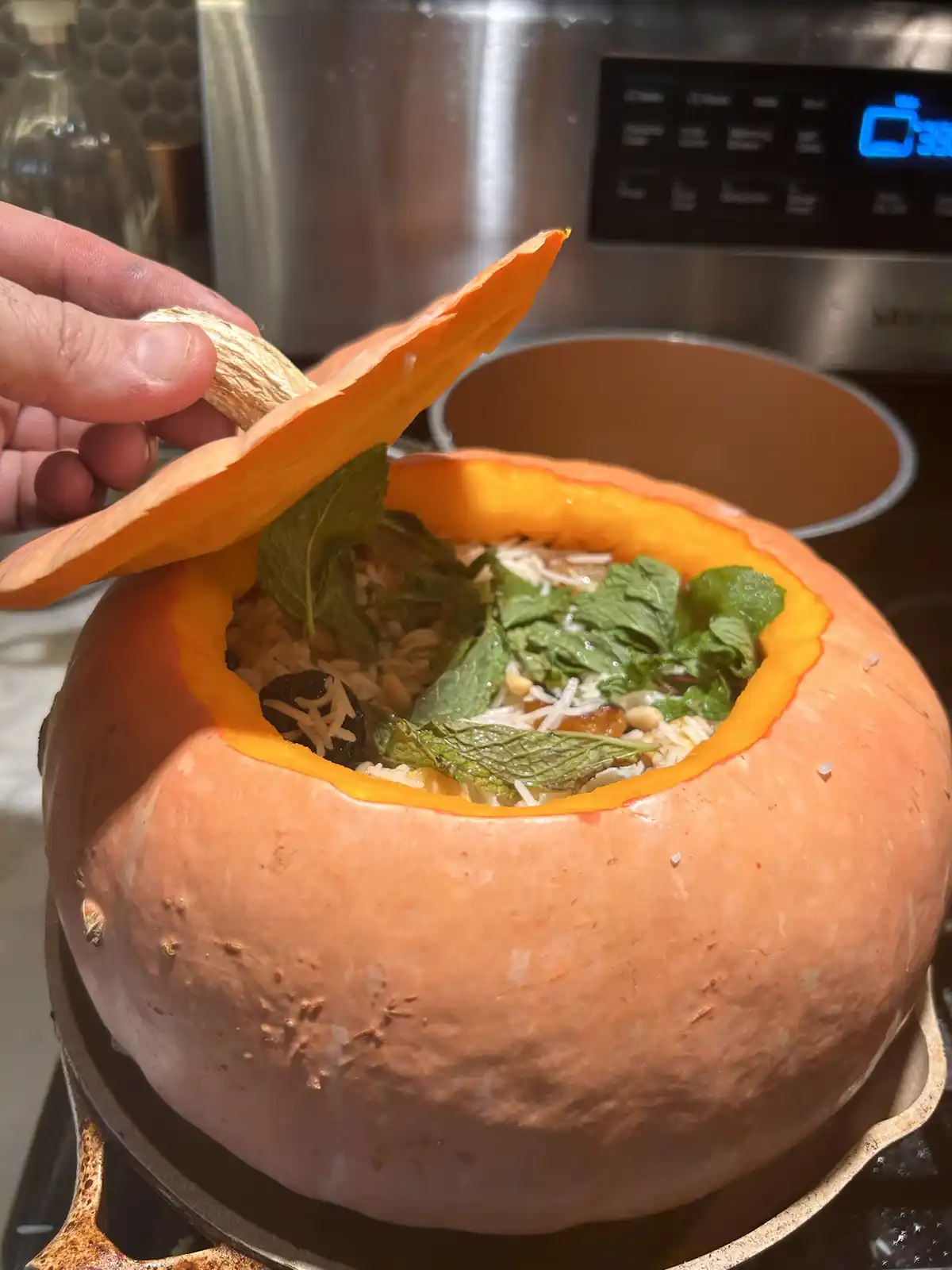 A hand lifts the lid of a hollowed-out orange pumpkin sitting on a stovetop, revealing a rice dish topped with mint leaves…