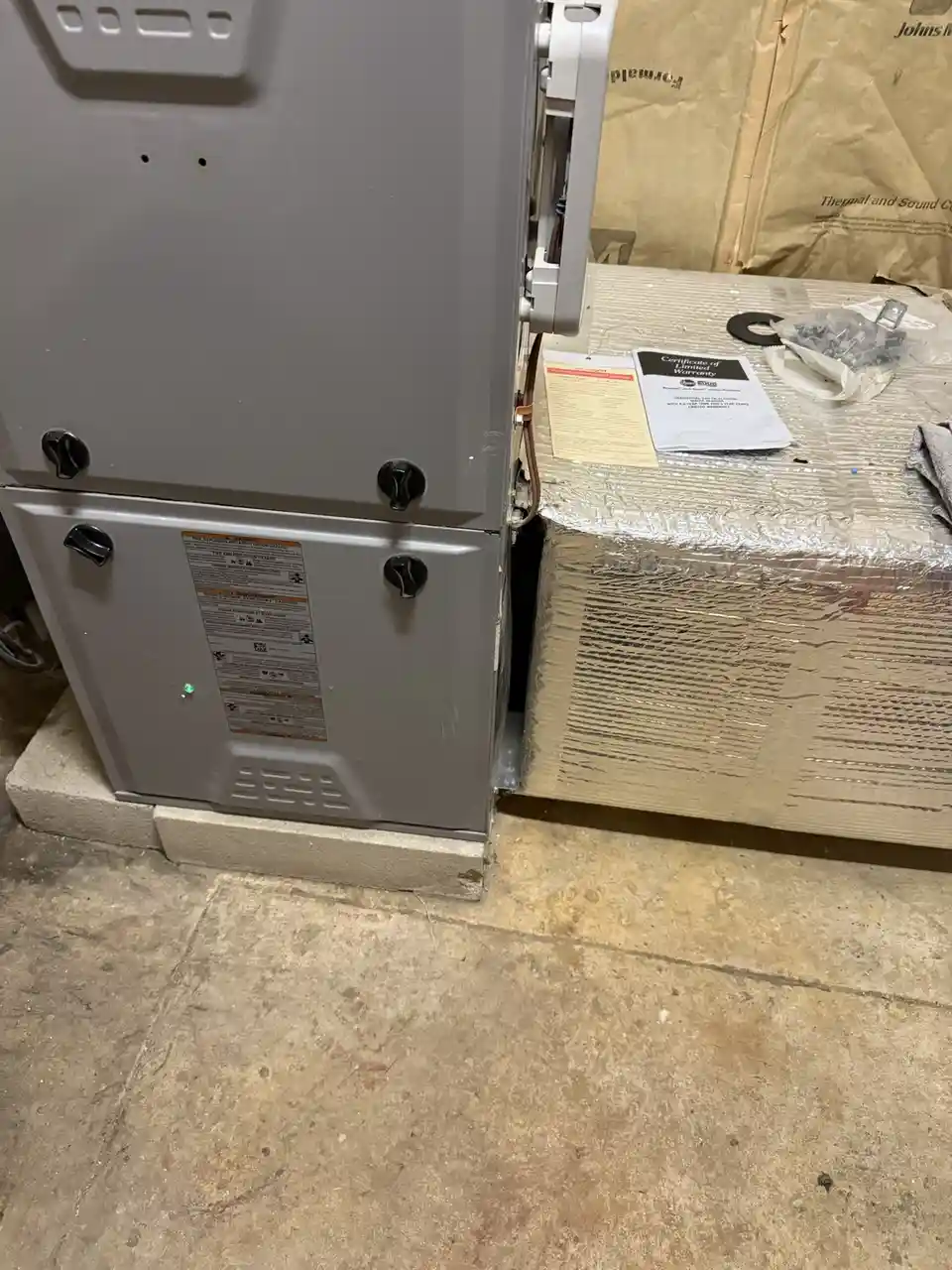 A gray water heater unit sits on a concrete slab next to a large, wrapped box, with a green light illuminated on its front…