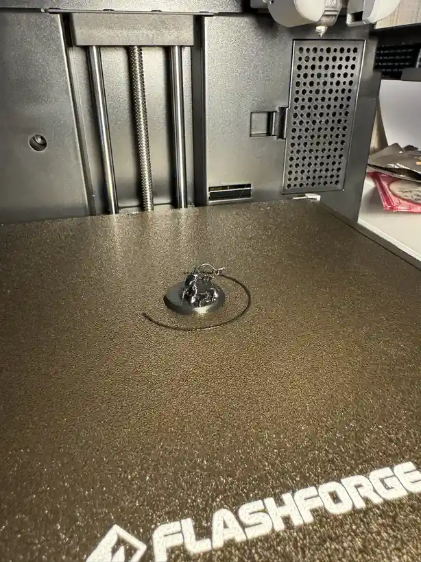 A close-up photo shows a small, shiny 3d-printed motorcycle figurine sitting on the build plate of a FlashForge 3d printer…