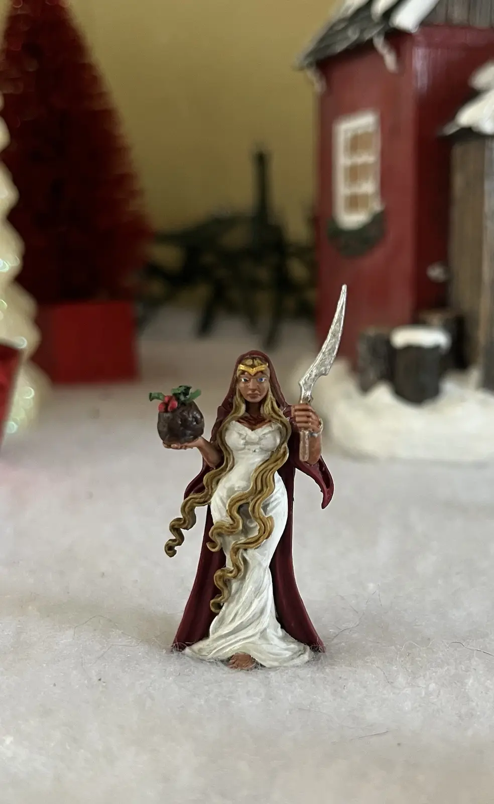 A figurine of a woman in a white dress and red cape stands in a snowy diorama with a red house and tree in the background…