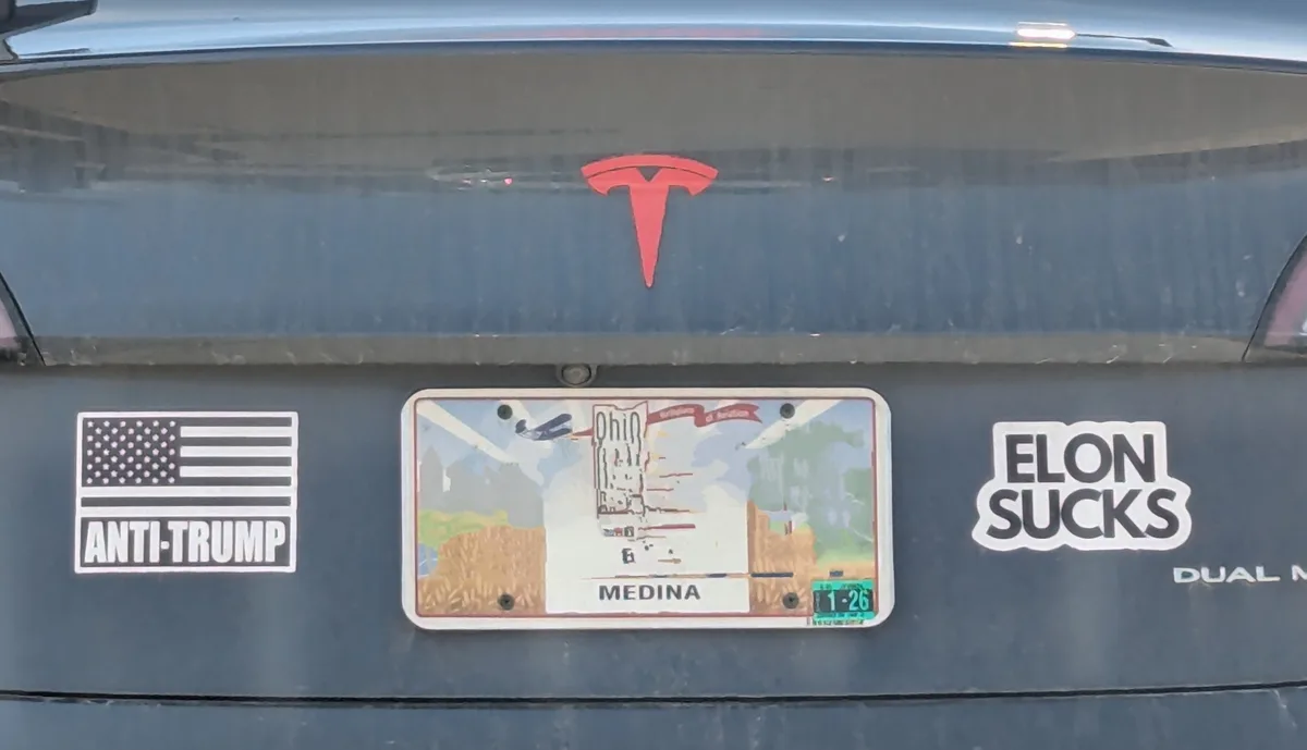 The rear of a dark-colored Tesla vehicle displays a red Tesla logo above an Ohio license plate reading 'MEDINA'. To the left…