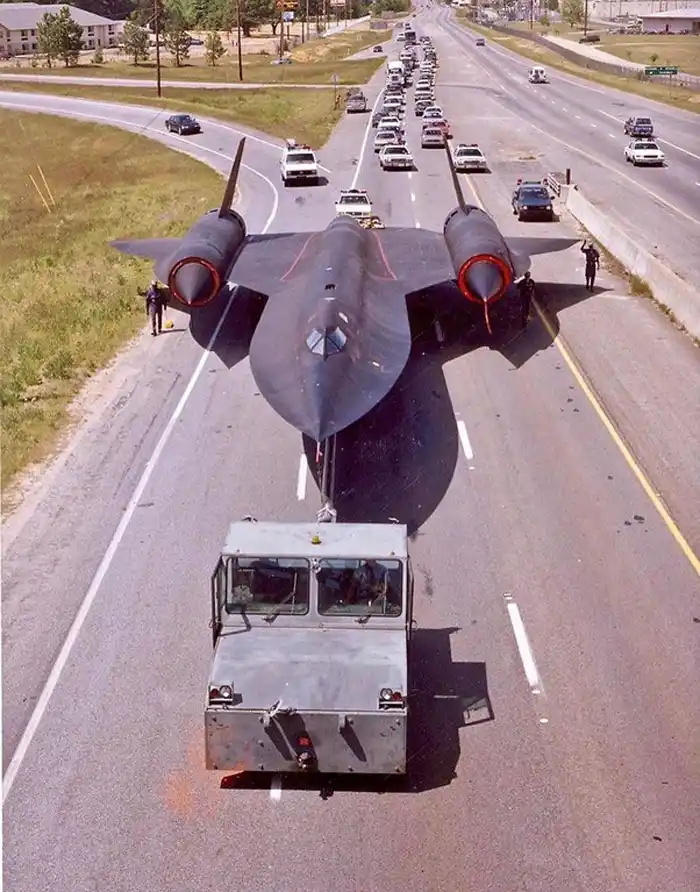 A black, delta-winged SR-71 Blackbird reconnaissance aircraft is being towed down a multi-lane highway by a small, boxy…