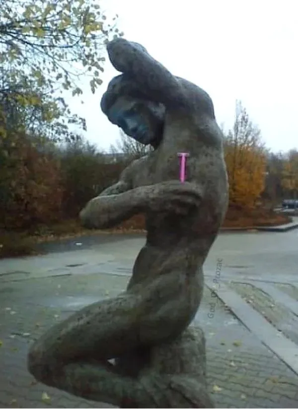A weathered outdoor statue of a muscular man in a crouching pose, with one hand on his head and the other on his chest, has…