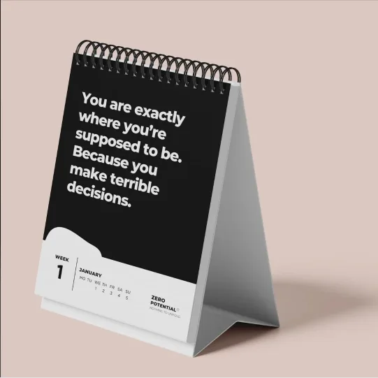 A black-and-white desk calendar with a spiral binding, standing upright on a stand, displays the text 'You are exactly where…