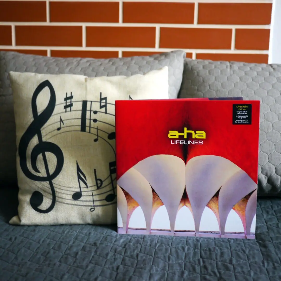 A red vinyl album cover for 'Lifelines' by a-ha sits on a gray couch next to a beige pillow with a large black treble clef…