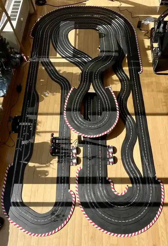An overhead shot shows a large, custom-built slot car racing track laid out on a light wood floor, shaped like the number 8…