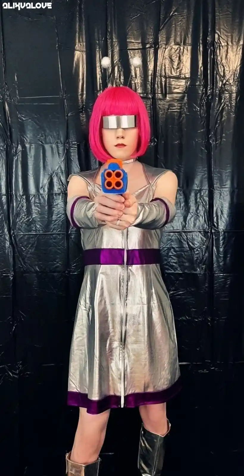 A person in a silver and purple alien costume with a pink bob wig and silver blindfold holds a blue and orange toy gun…