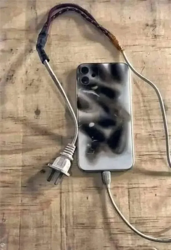 A smartphone with a visibly charred and melted back lies on a wooden surface, connected by a frayed, exposed electrical cord…