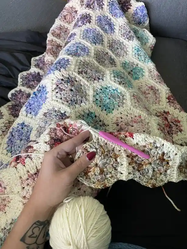 A hand with red nail polish and a tattoo holds a pink crochet hook while working on a large, colorful crocheted blanket made…
