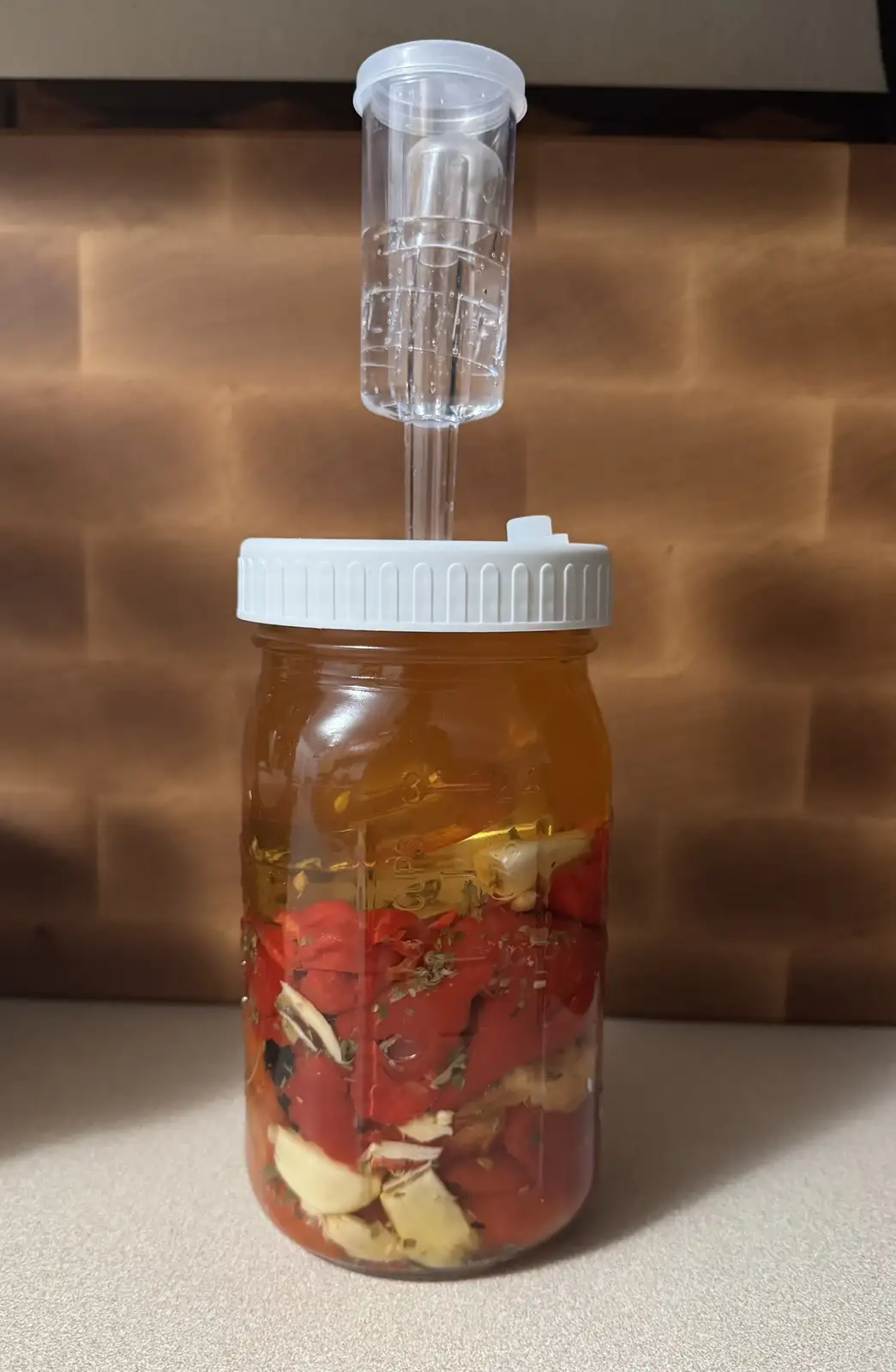 A glass mason jar filled with red peppers, garlic cloves, and herbs submerged in an amber liquid, topped with a white lid…
