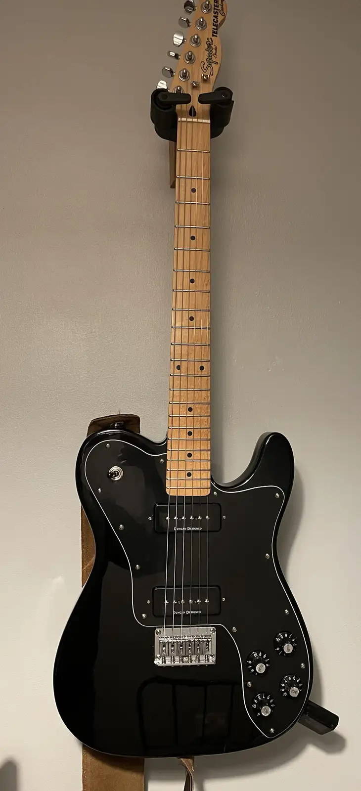 A black Squier Telecaster electric guitar with a maple neck and white binding is mounted vertically on a wall, held by a…